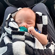 Kyzer is registered to the contest to win money with this photo: baby, sleeping, pacifier, blanket, checkered_pattern, infant, car_interior, lap, clothing, hand, peaceful, cozy, soft_texture, person, seat, steering_wheel, relaxation, child, portrait, closeup