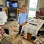 carpet, child, clothing, couch, cup, curtains, floor, indoors, living_room, person, remote_control, rug, shoes, socks, table, television, toddler, toy, window, wooden_furniture