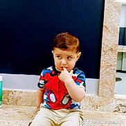 Salman is registered to the contest to win money with this photo: child, toddler, boy, portrait, sitting, stone_ledge, indoor, spiderman_shirt, superhero, colorful_clothing, shorts, sneakers, brown_hair, side_glance, finger_in_mouth, serious_expression, dark_background, marble, watermark, photography