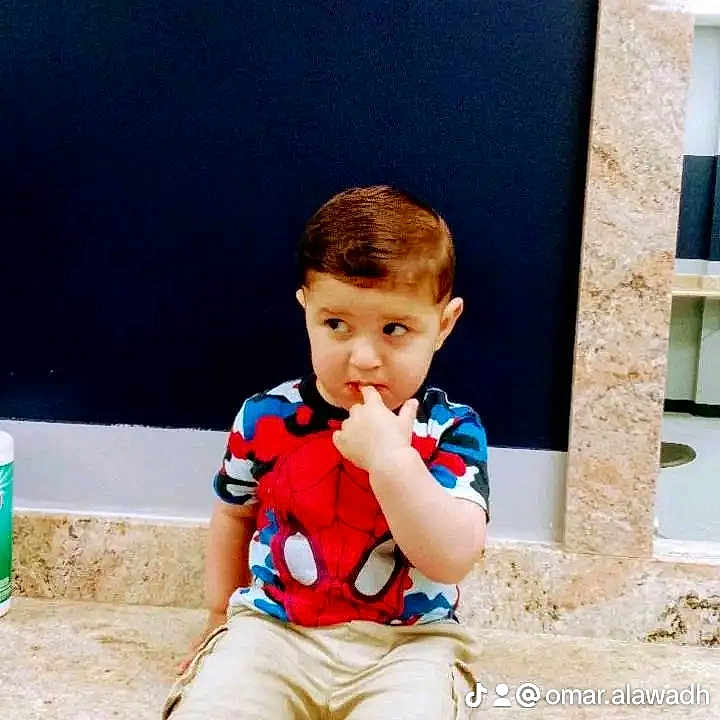 Salman is registered to the contest to win money with this photo: child, toddler, boy, portrait, sitting, stone_ledge, indoor, spiderman_shirt, superhero, colorful_clothing, shorts, sneakers, brown_hair, side_glance, finger_in_mouth, serious_expression, dark_background, marble, watermark, photography