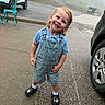 toddler, child, smiling, overalls, blue_shirt, crocs, outdoor, foggy, car, parking_lot, sidewalk, chair, happy, person, casual_clothing, shorts, daytime, head_tilt, cute, standing
