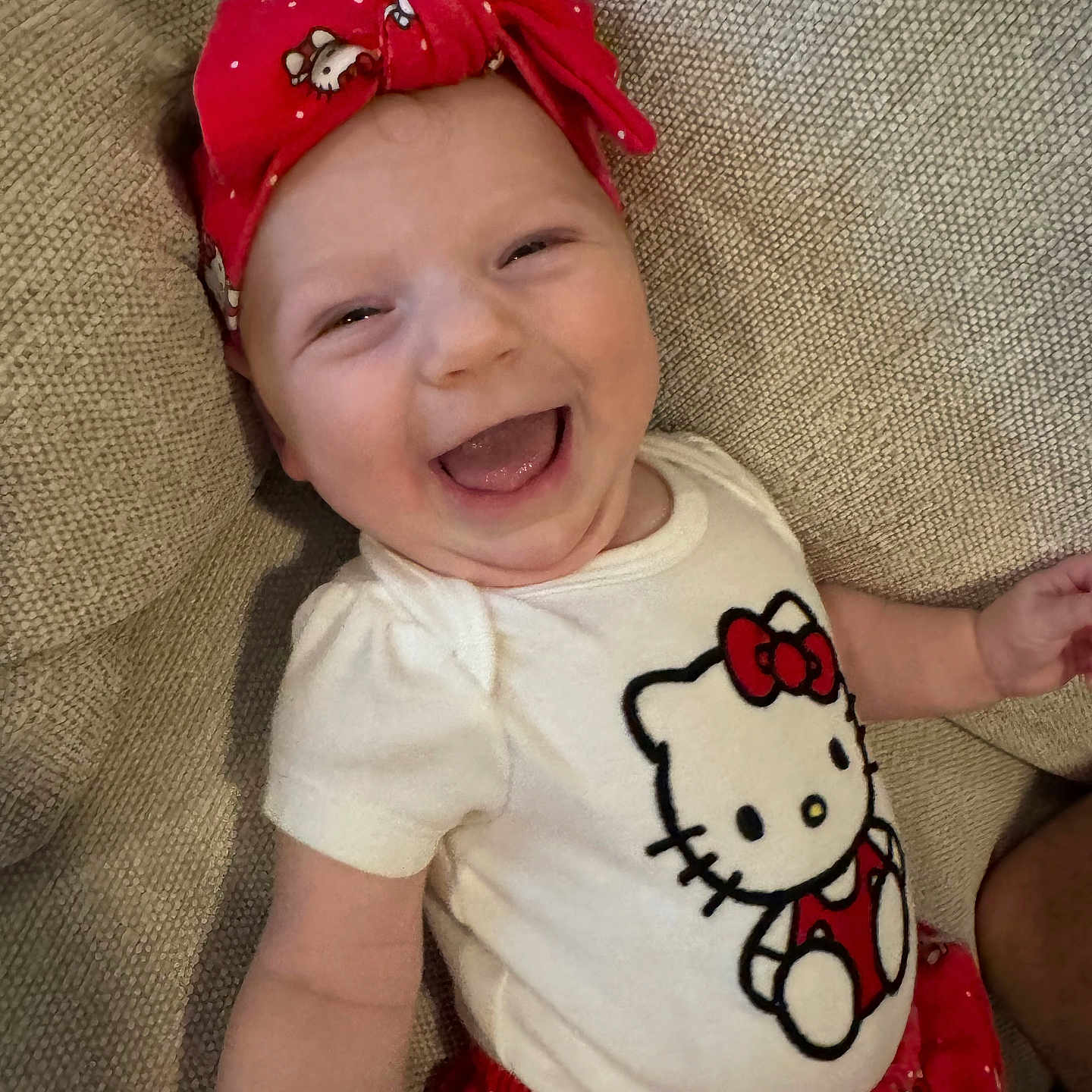 Malia joined the competition — help win amazing prizes! baby, child, smiling, laughing, headband, red_headband, hello_kitty, white_shirt, red_shorts, couch, beige_couch, happy, cute, infant, person, indoor, portrait, clothing, fabric, head