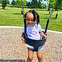 toddler, child, swing, playground, park, outdoor, grass, trees, sunny, daytime, smiling, braided_hair, beads, white_tshirt, denim_shorts, sneakers, happy, wood_chips, recreation, fun