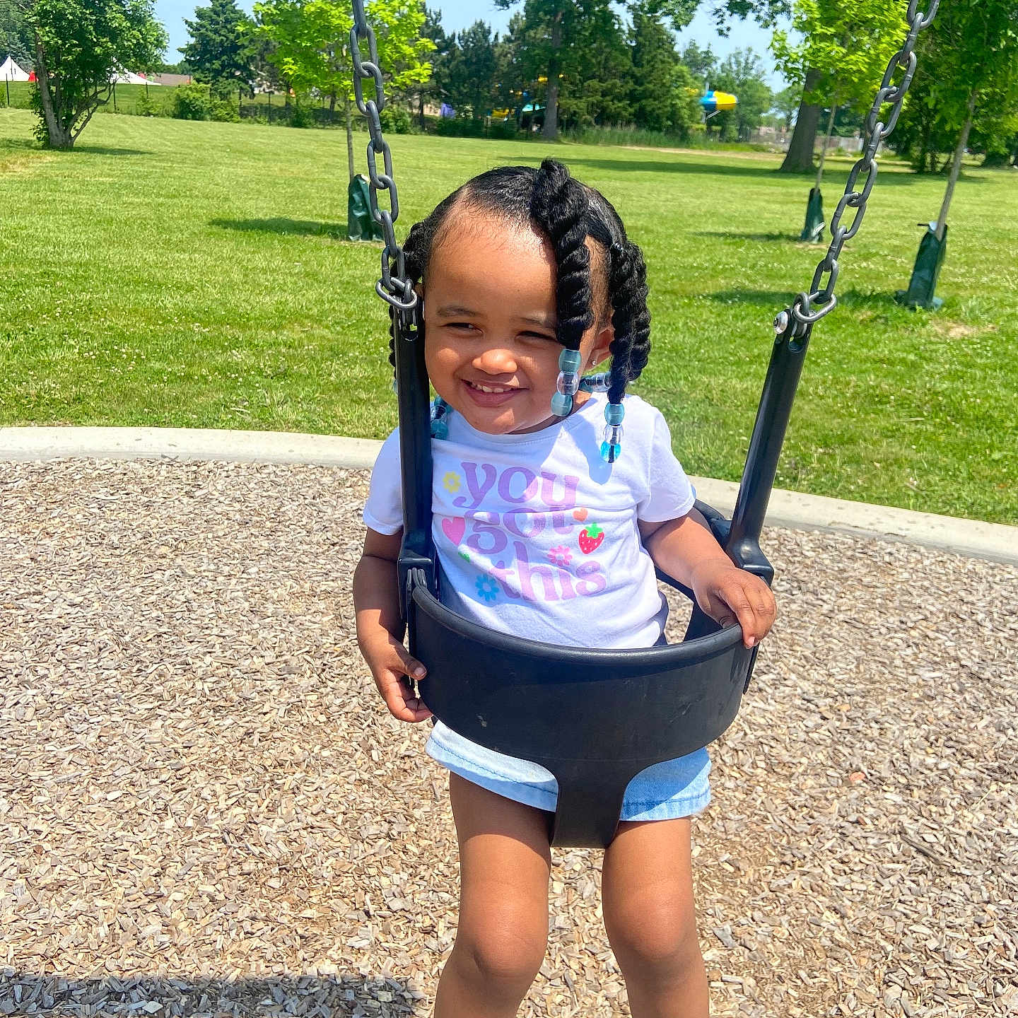 Skiley is registered to the contest to win money with this photo: beads, braided_hair, child, daytime, denim_shorts, fun, grass, happy, outdoor, park, playground, recreation, smiling, sneakers, sunny, swing, toddler, trees, white_tshirt, wood_chips