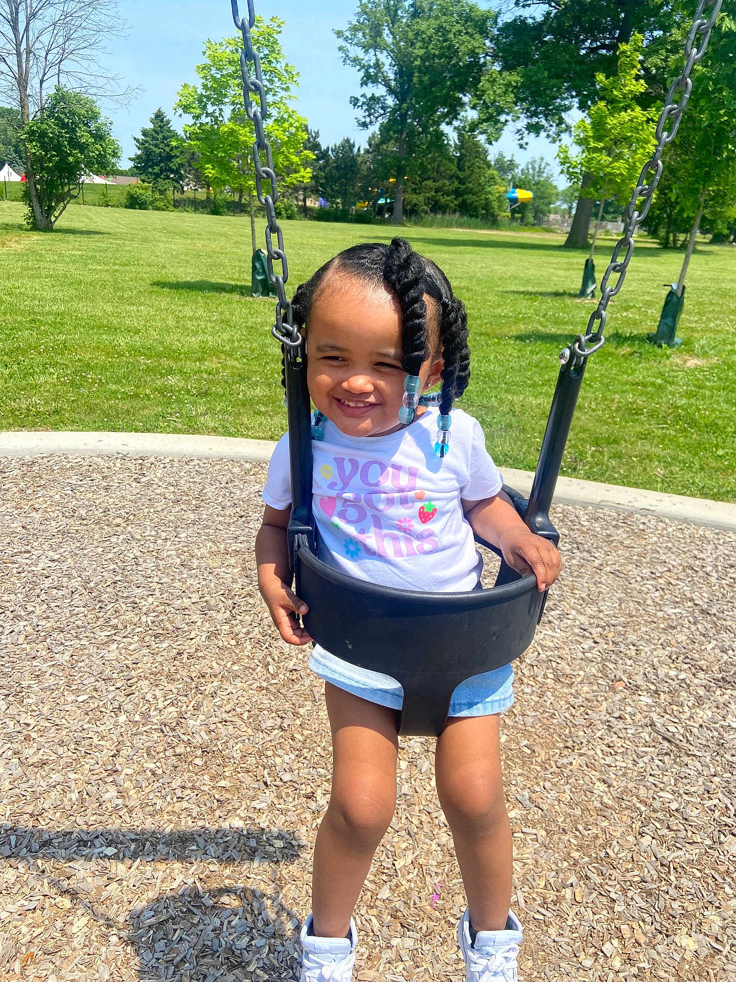 Skiley is registered to the contest to win money with this photo: toddler, child, swing, playground, park, outdoor, grass, trees, sunny, daytime, smiling, braided_hair, beads, white_tshirt, denim_shorts, sneakers, happy, wood_chips, recreation, fun