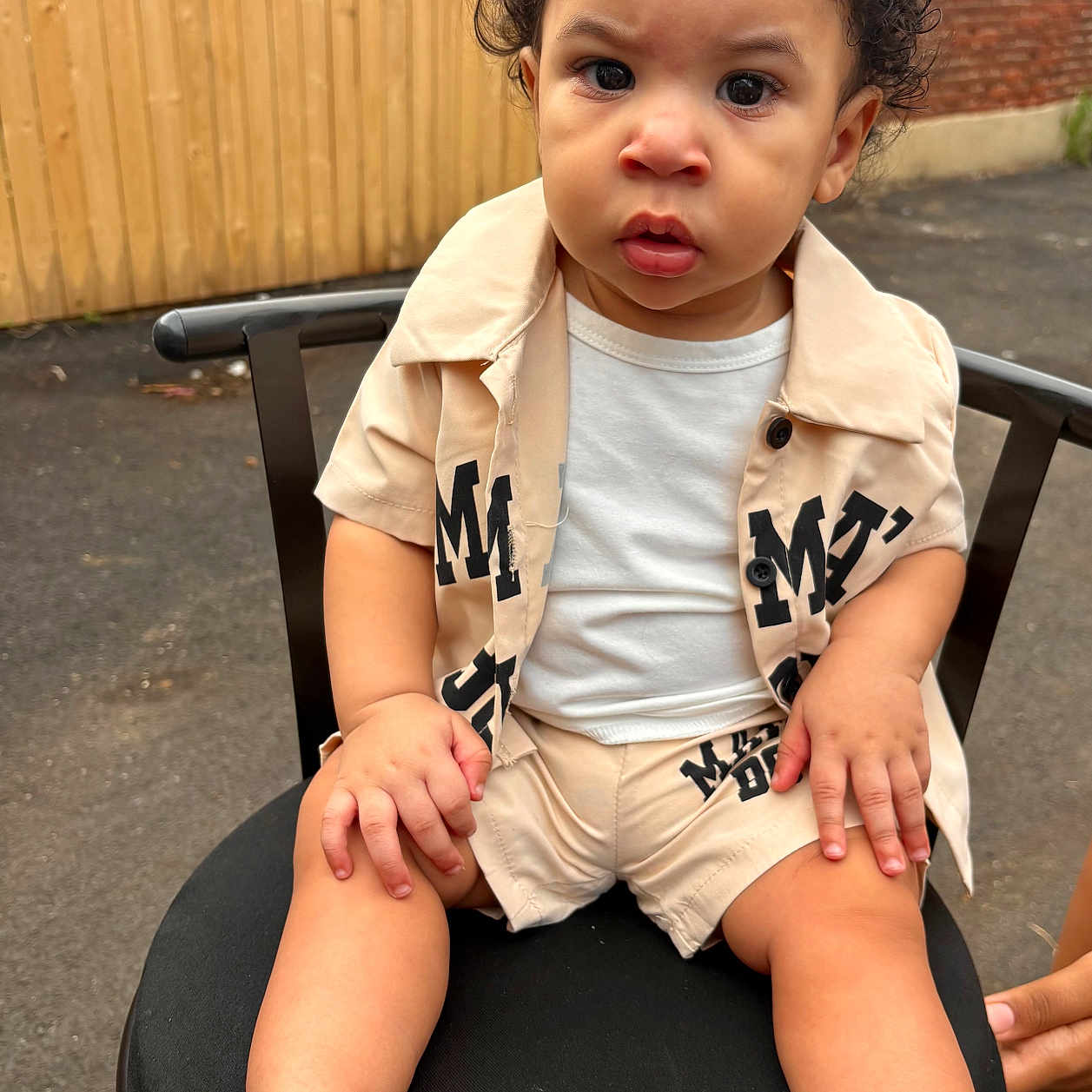 Jareth is registered to the contest to win money with this photo: toddler, child, curly_hair, beige_clothing, sandals, chair, outdoor, fence, brick_wall, serious_expression, sitting, person, young_child, shorts, shirt, footwear, skin, face, hands, summer_clothing
