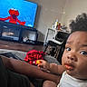 child, toddler, snack, food, face, messy, television, elmo, cartoon, living_room, couch, person, hair, curly_hair, clothing, hand, foot, red_cloth, table, chair