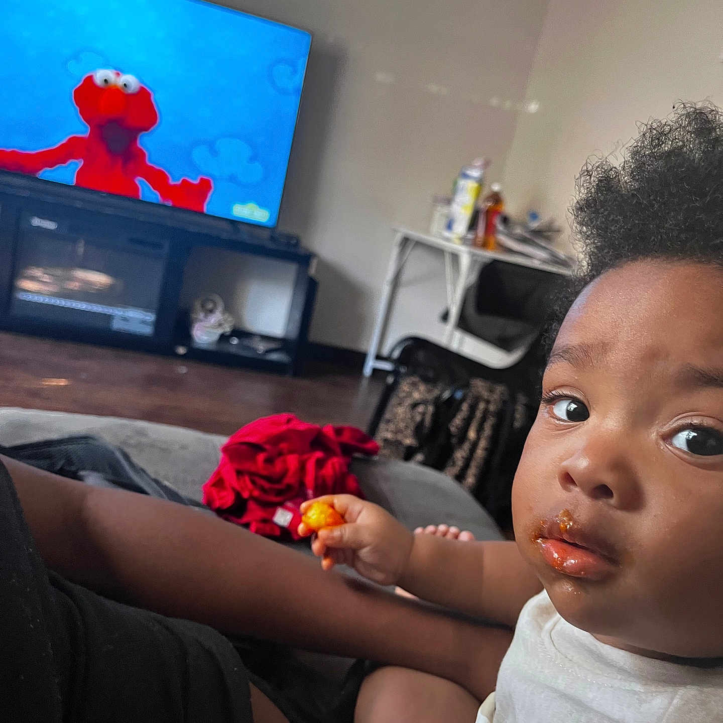 Kahjeer joined the competition — help win amazing prizes! child, toddler, snack, food, face, messy, television, elmo, cartoon, living_room, couch, person, hair, curly_hair, clothing, hand, foot, red_cloth, table, chair