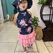 Elsa participe au concours pour gagner de l'argent avec cette photo : child, clothing, cute, dress, fashion, floral, footwear, furniture, girl, happy, hat, indoor, person, pink_skirt, plant, playful, pose, sandals, smiling, tile_floor