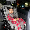 child, toddler, car_seat, car_interior, seatbelt, red_clothing, checkered_pattern, hair_buns, indoor, parking_garage, window, seat, young_child, casual_clothing, socks, curious_expression, vehicle, safety, infant, portrait