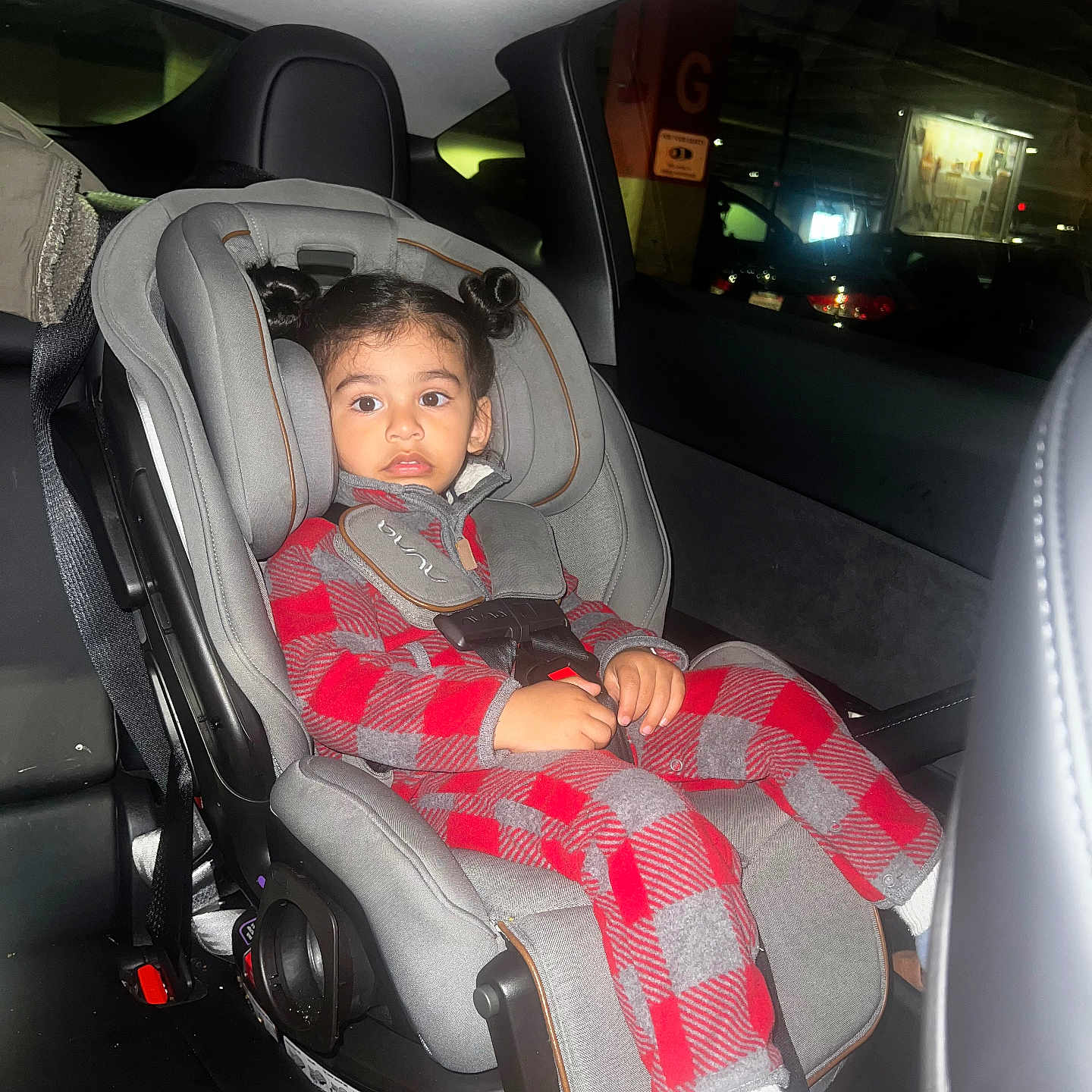 Soham is registered to the contest to win money with this photo: child, toddler, car_seat, car_interior, seatbelt, red_clothing, checkered_pattern, hair_buns, indoor, parking_garage, window, seat, young_child, casual_clothing, socks, curious_expression, vehicle, safety, infant, portrait