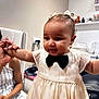 Anela joined the competition — help win amazing prizes! baby, child, dress, bow, earrings, indoor, person, hand, holding, cute, smiling, curly_hair, adult_hand, furniture, white, black_bow, hair_bows, face, happy, portrait