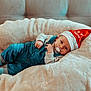 Arthur participe au concours pour gagner de l'argent avec cette photo : baby, blue_overalls, child, christmas_hat, clothing, comfort, cozy, cute, face, festive, holiday, indoor, infant, pacifier, person, plush_cushion, portrait, relaxing, socks, soft_texture