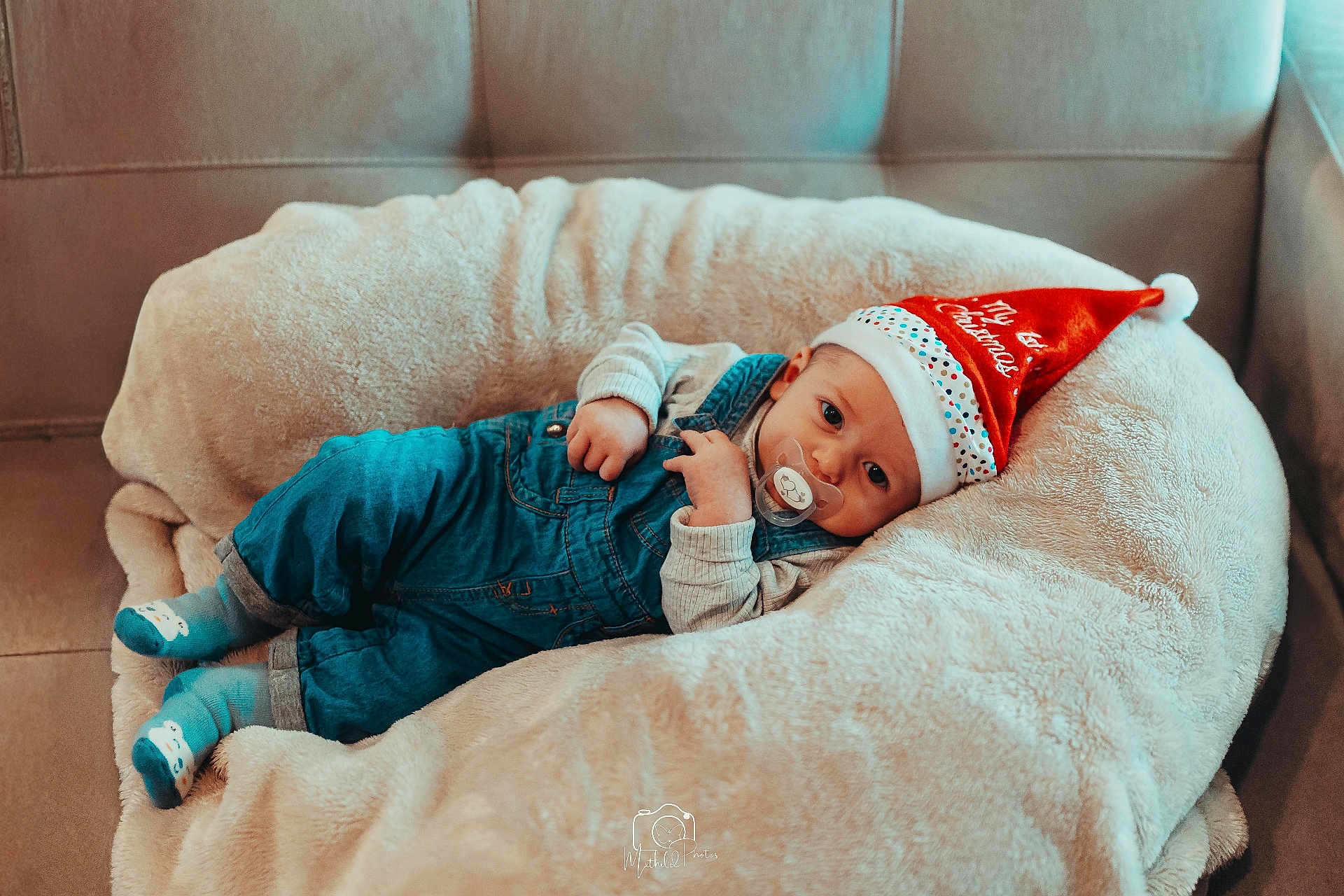 Arthur participe au concours pour gagner de l'argent avec cette photo : baby, blue_overalls, child, christmas_hat, clothing, comfort, cozy, cute, face, festive, holiday, indoor, infant, pacifier, person, plush_cushion, portrait, relaxing, socks, soft_texture