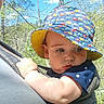 Jameson is registered to the contest to win money with this photo: baby, child, hat, bucket_hat, outdoor, nature, trees, sky, blue_sky, sunlight, carrier, person, infant, clothing, cute, expression, holding, forest, spring, daylight