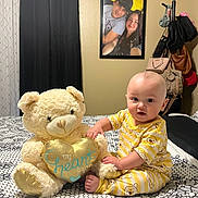 Eithan is registered to the contest to win money with this photo: baby, teddy_bear, stuffed_animal, bed, pajamas, yellow, heart, patterned_bedspread, portrait_photo, curtains, wall, room, furniture, clothing, child, indoor, smiling, person, coatrack, bags