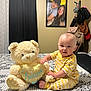 baby, teddy_bear, stuffed_animal, bed, pajamas, yellow, heart, patterned_bedspread, portrait_photo, curtains, wall, room, furniture, clothing, child, indoor, smiling, person, coatrack, bags