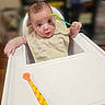 baby, high_chair, spoon, bib, infant, child, cute, feeding, table, face, hand, expression, indoor, person, curious, young, seat, food, kitchen, bright