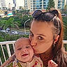 baby, woman, kiss, smile, outdoor, balcony, sunglasses, tattoo, long_hair, urban, trees, buildings, daylight, happy, person, child, affection, holding, face, casual