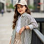 child, girl, urban, street_style, cap, striped_shirt, pleated_skirt, watch, leaning, railing, confident, smiling, long_hair, casual, fashion, young, outdoor, daylight, portrait, city