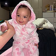Arleth joined the competition — help win amazing prizes! baby, child, pink_onesie, hood, smiling, indoor, bedroom, teddy_bear, dresser, blanket, hand, face, person, portrait, cute, infant, cozy, fuzzy_clothing, furniture, photo_frame