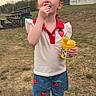 Brooklynn is registered to the contest to win money with this photo: child, red_hair, outdoor, grass, dirt, snack, yellow_container, strawberry, shorts, shirt, cloudy_sky, trees, building, casual, footwear, happy, person, summer_clothing, playing, daytime