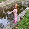 child, girl, red_hair, pink_clothing, water, canal, stone_wall, grass, leaves, autumn, outdoor, playful, smiling, park, reflection, trees, sidewalk, casual, nature, daytime