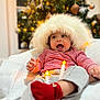 baby, child, smiling, hat, red_striped_shirt, red_socks, blanket, christmas_tree, ornaments, holiday, festive, indoor, cozy, happy, person, face, cute, warm, celebration, soft_lighting