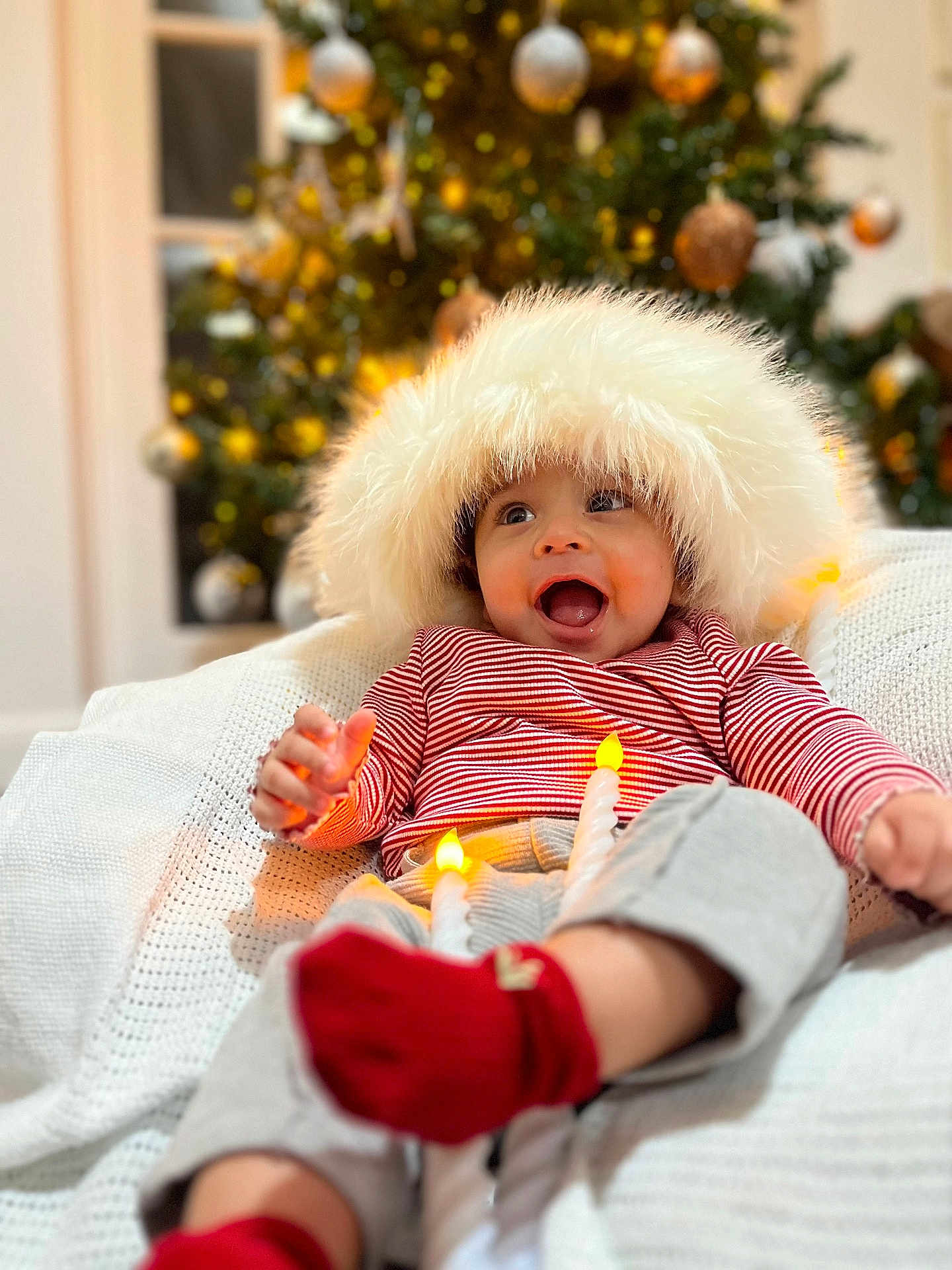 Jayden joined the competition — help win amazing prizes! baby, child, smiling, hat, red_striped_shirt, red_socks, blanket, christmas_tree, ornaments, holiday, festive, indoor, cozy, happy, person, face, cute, warm, celebration, soft_lighting