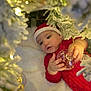 baby, infant, christmas, holiday, ornament, santa_hat, red_clothing, blanket, decorated_tree, snowy_branches, soft_lighting, curious, festive, indoors, child, face, hands, holiday_season, cute, toddler