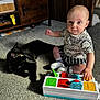 baby, child, cat, black_cat, toy, carpet, indoor, furniture, wooden_cabinet, colorful_toy, sitting, playing, curious, sunlight, shadow, striped_clothing, white_socks, person, floor, pet