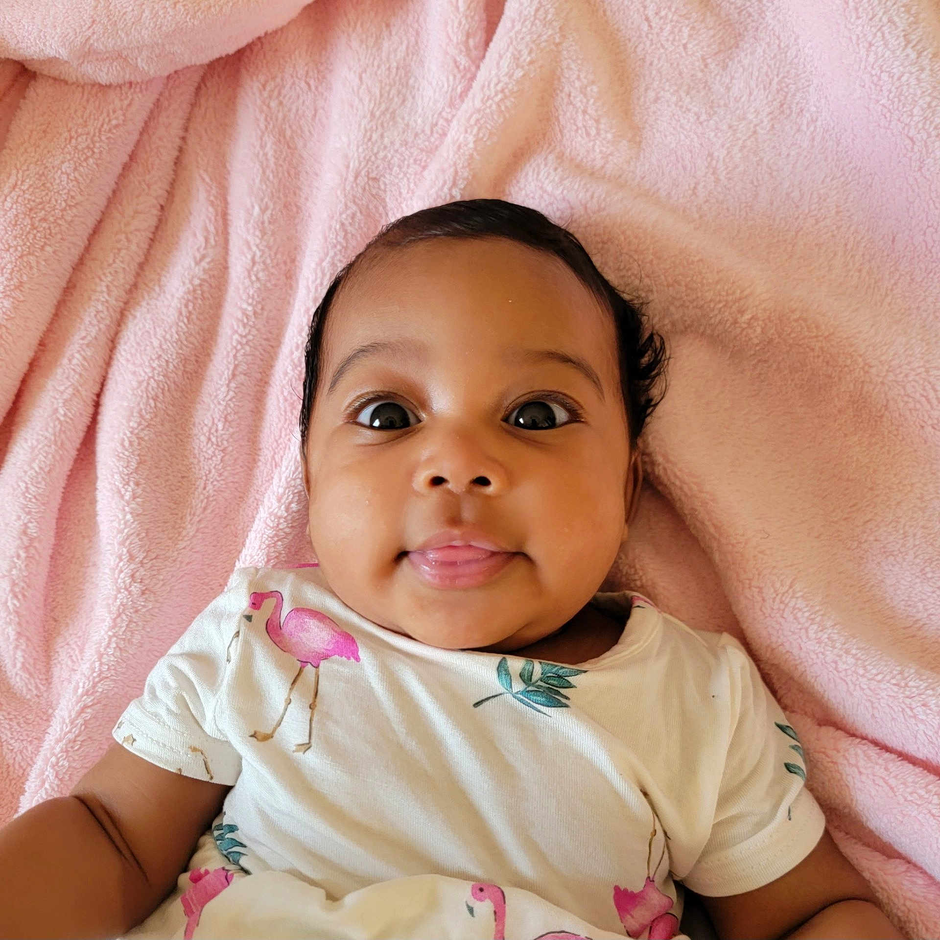 Ava joined the competition — help win amazing prizes! baby, infant, child, pink_blanket, flamingo_print, white_shirt, tongue_out, cute, lying_down, soft_texture, indoors, portrait, happy, curious, face, person, skin, head, eyes, smile
