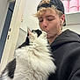 person, cat, indoor, black_hoodie, cap, kiss, fluffy_cat, white_black_cat, curly_hair, earbuds, shelf, toilet_paper, door, wall, facial_piercing, whiskers, affection, pet, close_up, casual