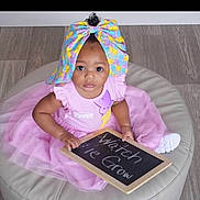 Haylyn is registered to the contest to win money with this photo: baby, child, pink_dress, tutu, headwrap, colorful_bow, chalkboard, floor, cushion, sitting, curious_eyes, indoor, person, cute, portrait, infant, hand, clothing, footwear, message