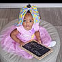 baby, child, pink_dress, tutu, headwrap, colorful_bow, chalkboard, floor, cushion, sitting, curious_eyes, indoor, person, cute, portrait, infant, hand, clothing, footwear, message