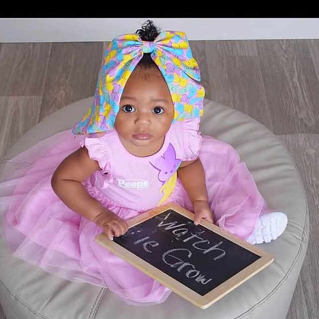 Haylyn is registered to the contest to win money with this photo: baby, chalkboard, child, clothing, colorful_bow, curious_eyes, cushion, cute, floor, footwear, hand, headwrap, indoor, infant, message, person, pink_dress, portrait, sitting, tutu