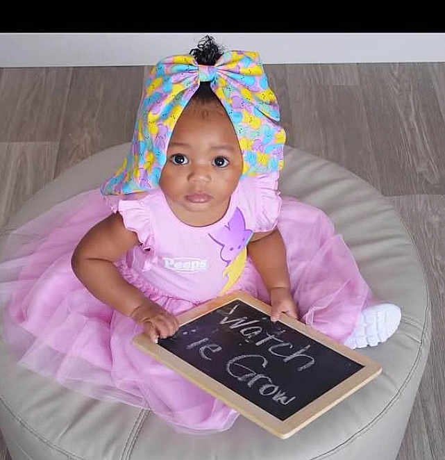 Haylyn is registered to the contest to win money with this photo: baby, child, pink_dress, tutu, headwrap, colorful_bow, chalkboard, floor, cushion, sitting, curious_eyes, indoor, person, cute, portrait, infant, hand, clothing, footwear, message