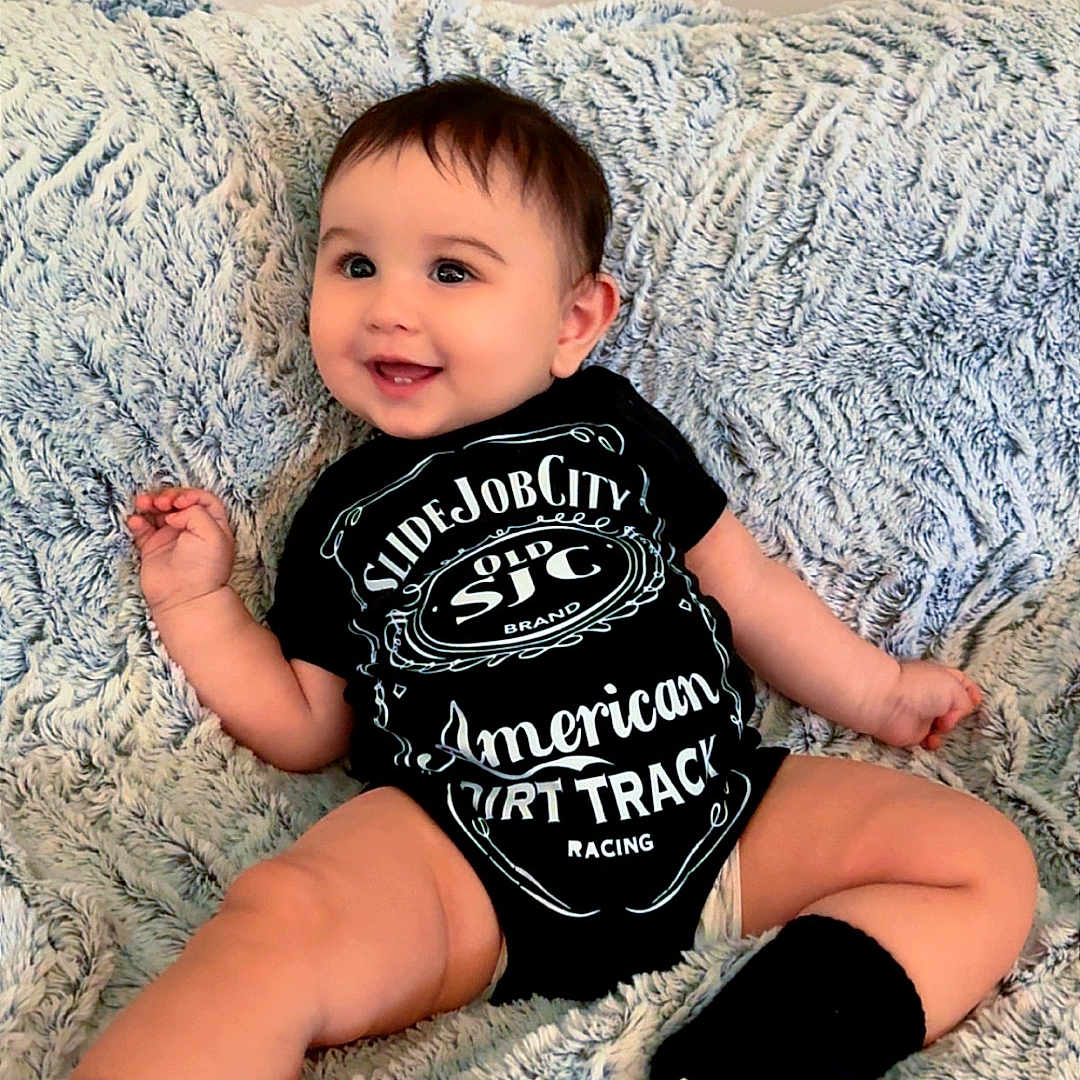Cruz joined the competition — help win amazing prizes! baby, smiling, onesie, black_clothing, sitting, blanket, indoor, cute, infant, child, happy, face, person, feet, soft_texture, cozy, portrait, young_child, sock, fuzzy