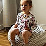 child, toddler, indoor, knit_pouf, toy, pink, mickey_mouse, minnie_mouse, clothing, socks, floor, wooden_floor, curtain, earrings, necklace, diaper, expression, portrait, sitting, home