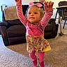 toddler, child, jumping, pink_shoes, striped_shirt, colorful_dress, living_room, carpet, couch, high_chair, smiling, happy, arms_up, bow, curly_hair, indoor, furniture, portrait, person, playful