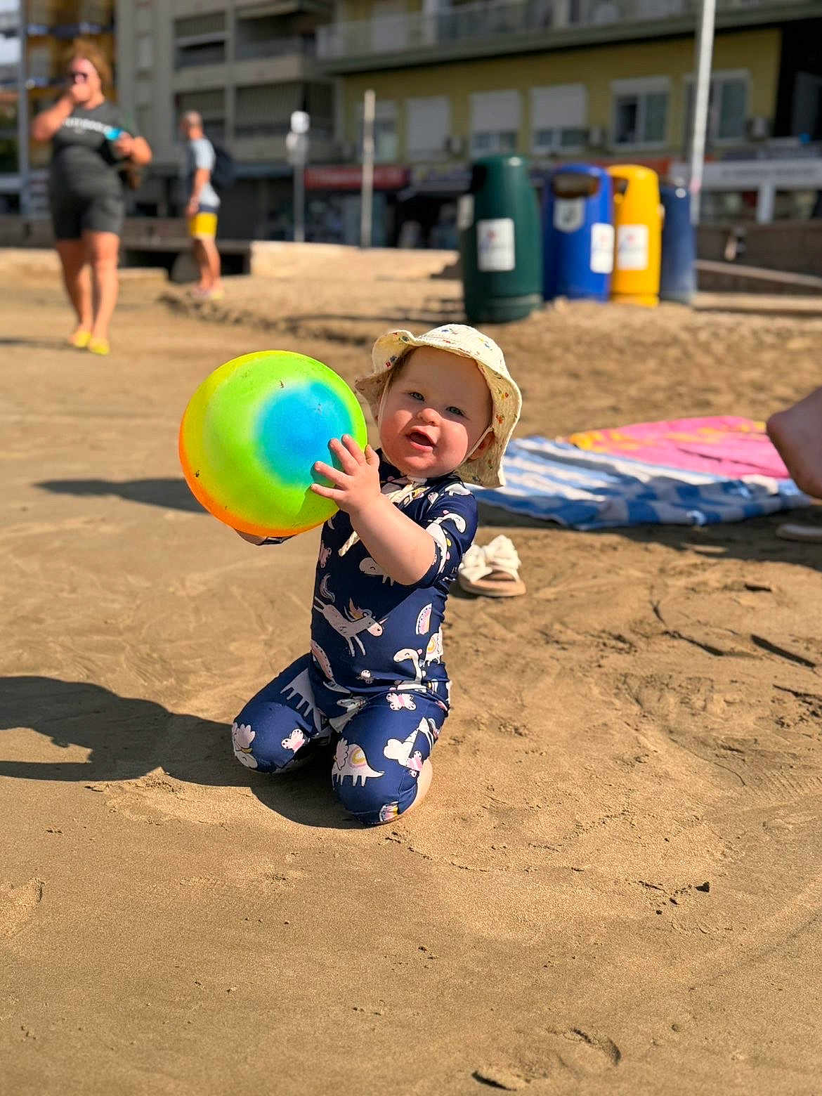 Summer is registered to the contest to win money with this photo: toddler, child, beach, sand, ball, hat, sun_hat, colorful, play, outdoor, smiling, kneeling, sunny, summer, footwear, background_people, recycling_bins, casual_clothing, daylight, happy