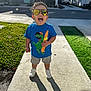 toddler, child, sunglasses, blue_tshirt, dinosaur_graphic, shorts, white_shoes, sidewalk, shadow, grass, suburban, house, car, sunlight, happy, smiling, outdoor, daytime, person, curly_hair
