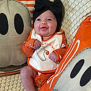 Brayleigh is registered to the contest to win money with this photo: baby, infant, smiling, black_bow, orange_clothing, halloween, ghost_pillow, decorative_pillow, blanket, couch, happy, cute, indoor, portrait, child, headwear, holiday, festive, infant_clothing, cozy
