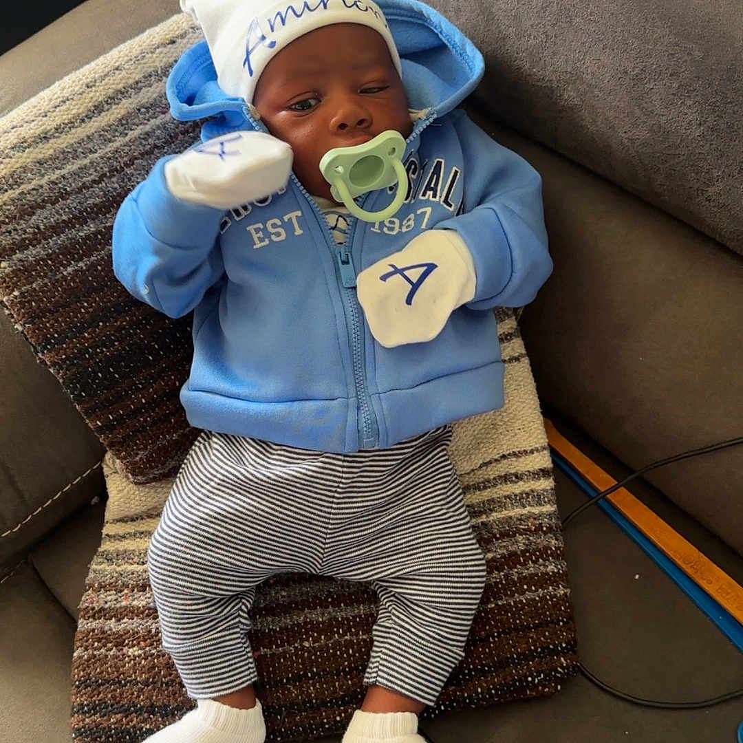 Amirion is registered to the contest to win money with this photo: baby, blue_hoodie, child, couch, cute, face, hat, home, indoor, infant, looking, mittens, pacifier, pillow, relaxed, seated, soft_lighting, striped_pants, white_socks, young