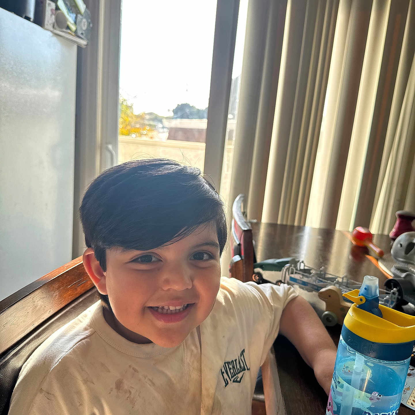 Andrew Matteo Romero is registered to the contest to win money with this photo: boy, casual_clothing, chair, child, curtain, daylight, face, happy, home, indoor, person, playtime, shirt, smile, sunlight, table, toy, water_bottle, window, wooden_furniture