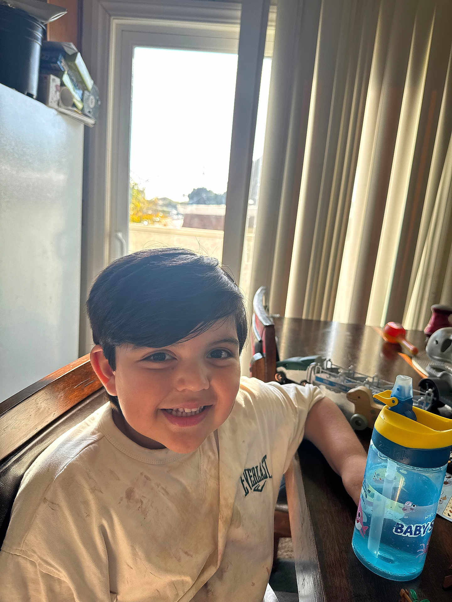 Andrew Matteo Romero is registered to the contest to win money with this photo: child, boy, smile, indoor, table, chair, toy, water_bottle, window, curtain, shirt, wooden_furniture, sunlight, happy, face, person, casual_clothing, home, playtime, daylight