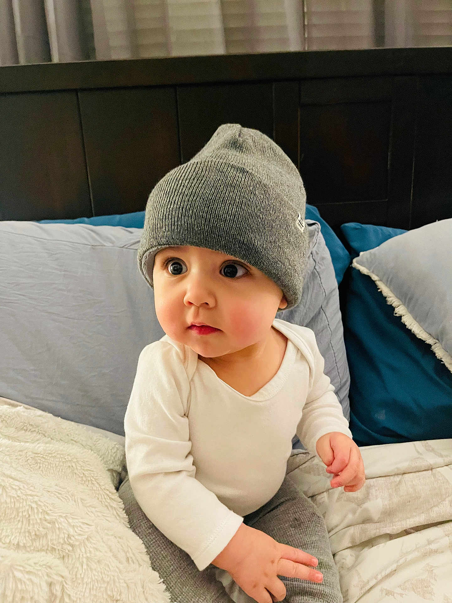 Isaac is registered to the contest to win money with this photo: baby, child, beanie, hat, white_clothing, gray_clothing, bed, pillow, blanket, indoor, curious, cute, infant, portrait, sitting, soft_lighting, headwear, couch, young_child, person