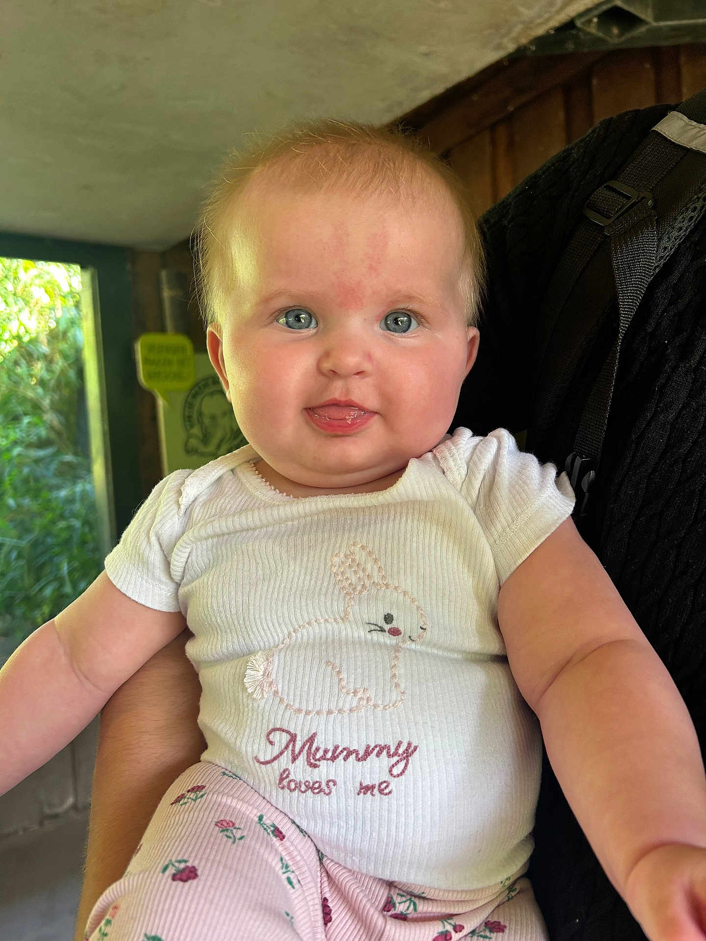 Laura joined the competition — help win amazing prizes! baby, child, infant, face, blue_eyes, white_shirt, bunny_design, text_on_clothing, pink_pants, floral_pattern, person, indoor, window, greenery, cute, smiling, holding, skin, tongue, short_hair