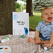 Wyatt is registered to the contest to win money with this photo: baby, child, outdoor, grass, tree, paint, paintbrush, bottle, blanket, smile, happy, celebration, sign, half_birthday, blue_shirt, shorts, tooth, face, sitting, nature