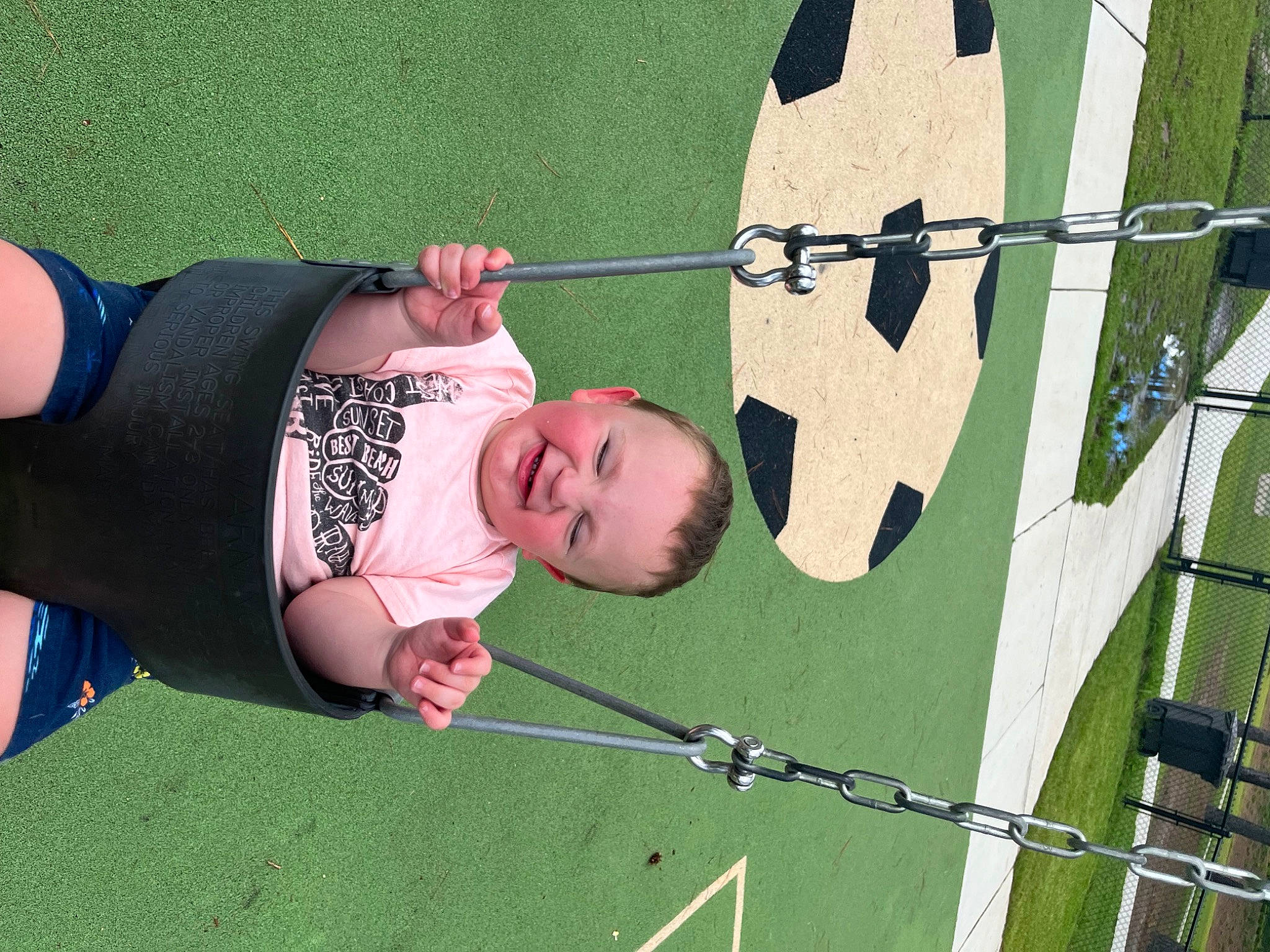 Hayden is registered to the contest to win money with this photo: baby, child, eye, fence, fun, grass, happy, hat, human_body, leaf, leisure, outdoor_play_equipment, people_in_nature, person, plant, play, recreation, sitting, swing, toddler